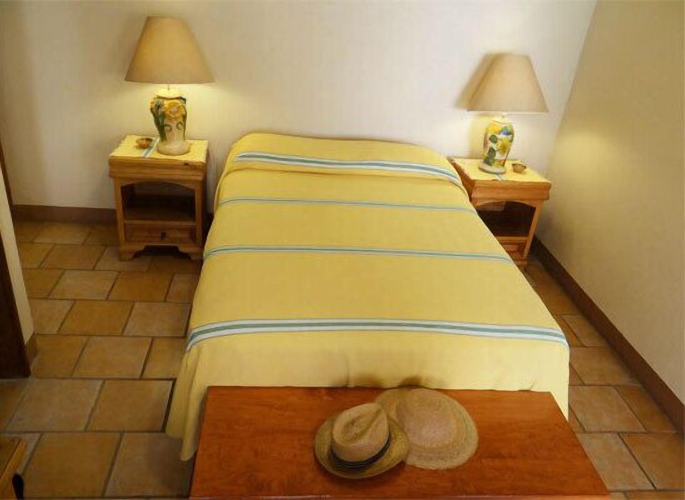 Une chambre d’hôtel avec un lit, deux tables de chevet, une lampe sur chaque table, une petite table en bois et un chapeau posé sur une autre table en bois.