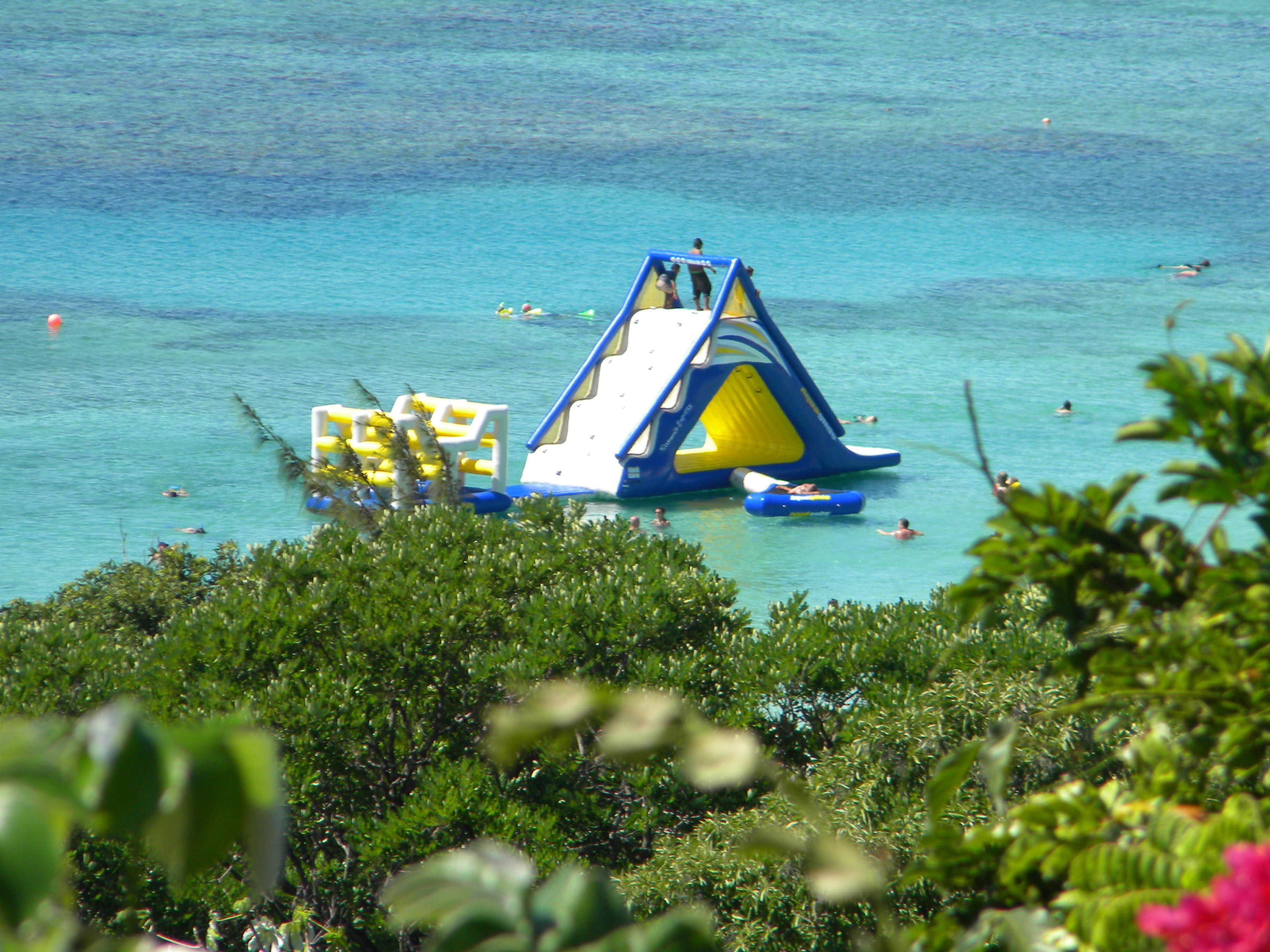 A water play area with slides and inflatable structures in a clear blue pool, surrounded by greenery.