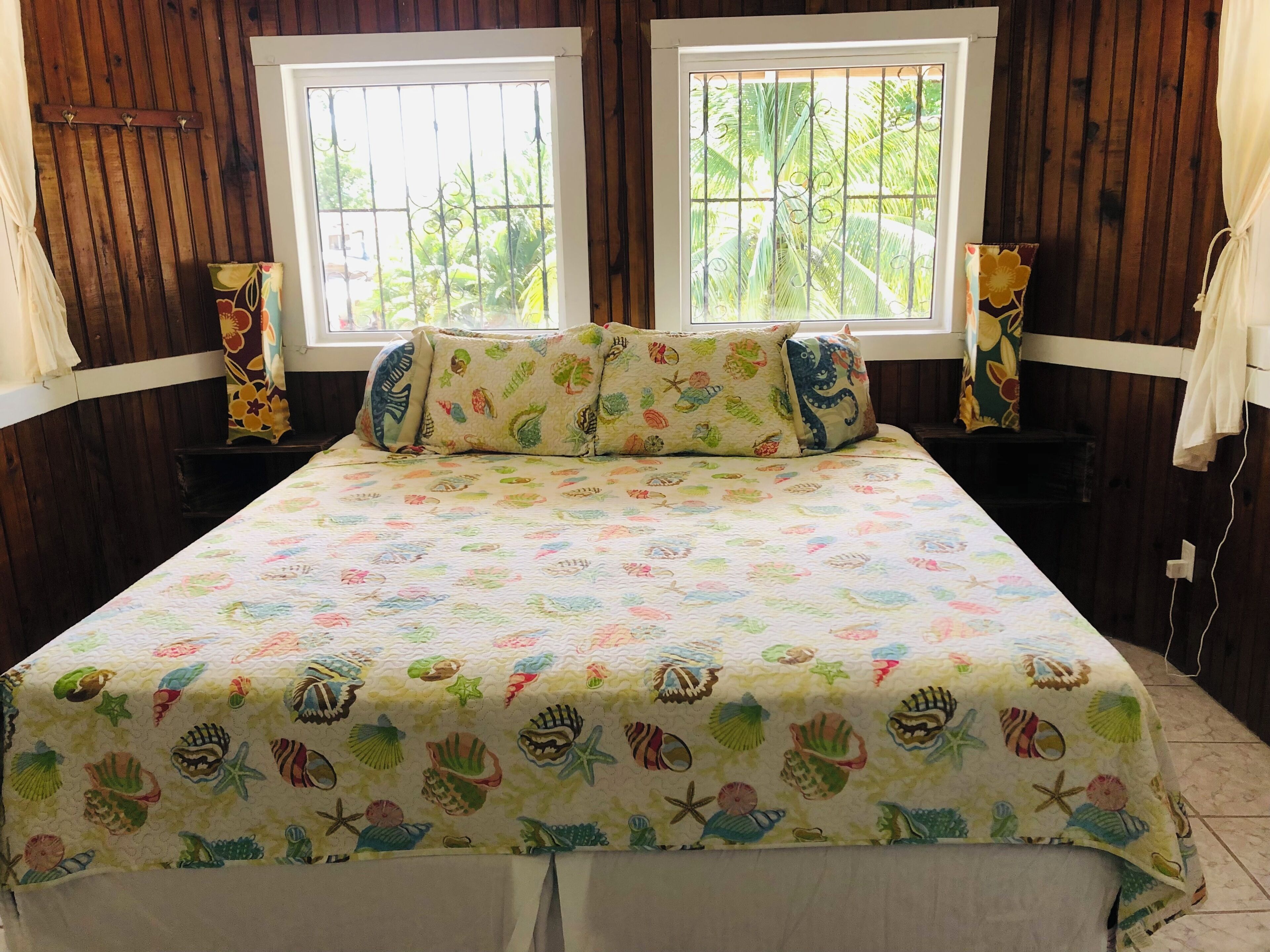 A bed with a colorful seashell and starfish bedspread, two windows with metal bars, wooden walls, and a curtain on the left.