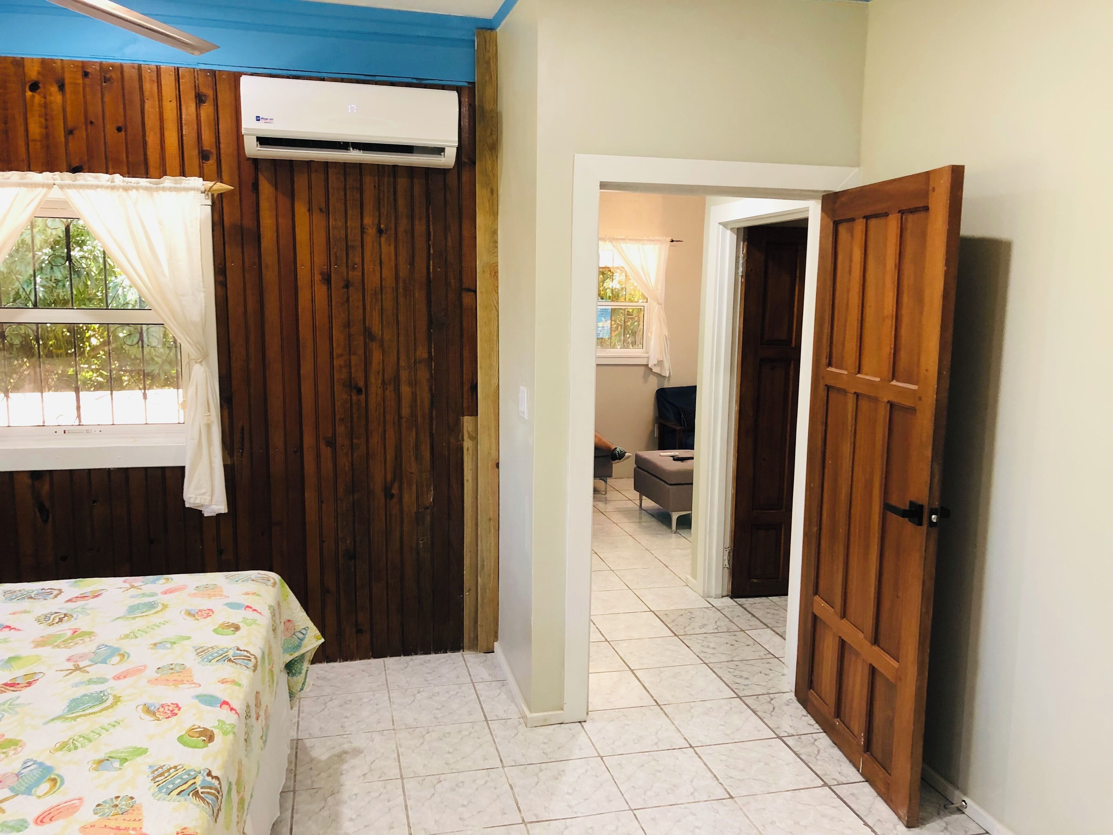 Een slaapkamer met een bed, een airconditioner, een raam met gordijnen, en een houten deur die leidt naar een andere kamer.