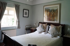 A bedroom with a bed, a window with curtains, a radiator, a bedside table with a lamp, and a framed painting on the wall.