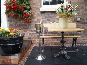 A brick wall with a window, a metal bucket, a wooden table with a bench, and a potted plant with red flowers.