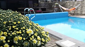Une piscine dont le bord est bleu, entourée de fleurs jaunes et blanches, et avec des parasols rouges en arrière-plan.