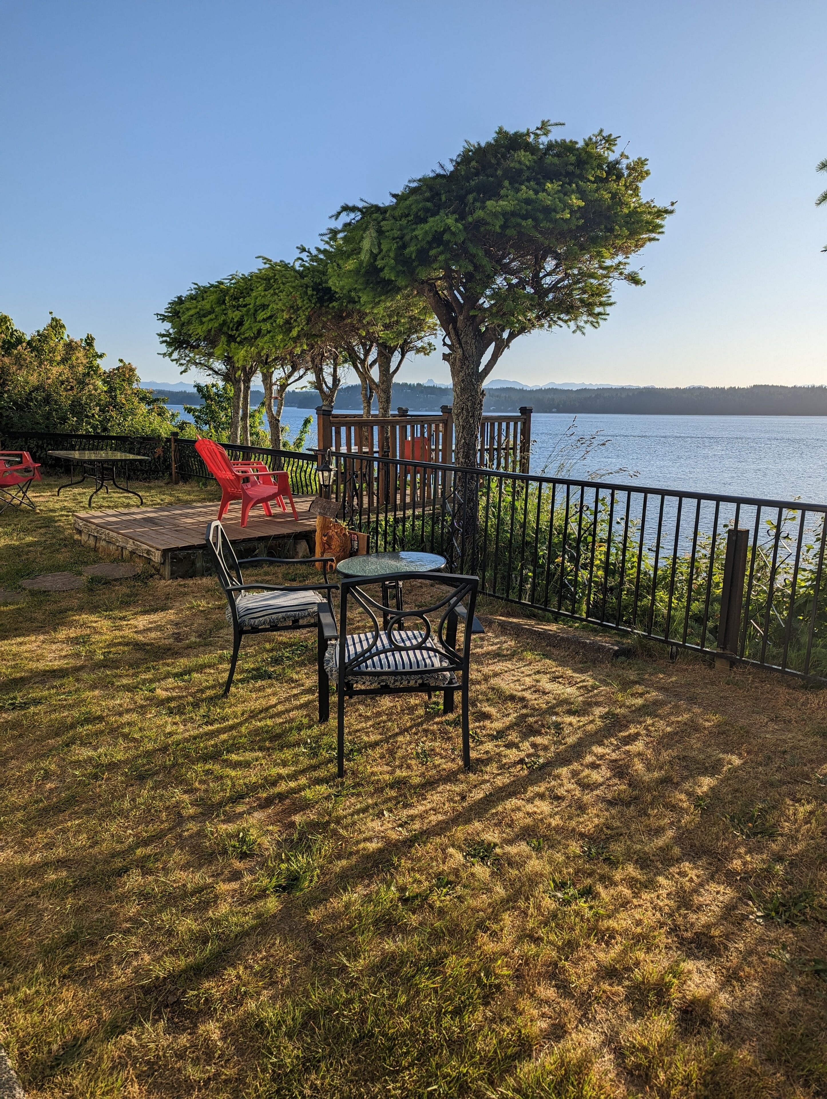 An outdoor patio with a table and chairs, overlooking a lake with trees and a wooden deck.