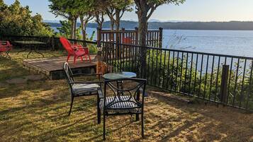 An outdoor patio with a table and chairs, overlooking a lake with trees and a wooden deck.