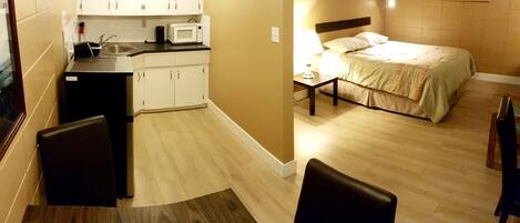 Habitación de hotel pequeña con kitchenette, cama, mesita de noche con lámpara, y zona de comedor con mesa y sillas.