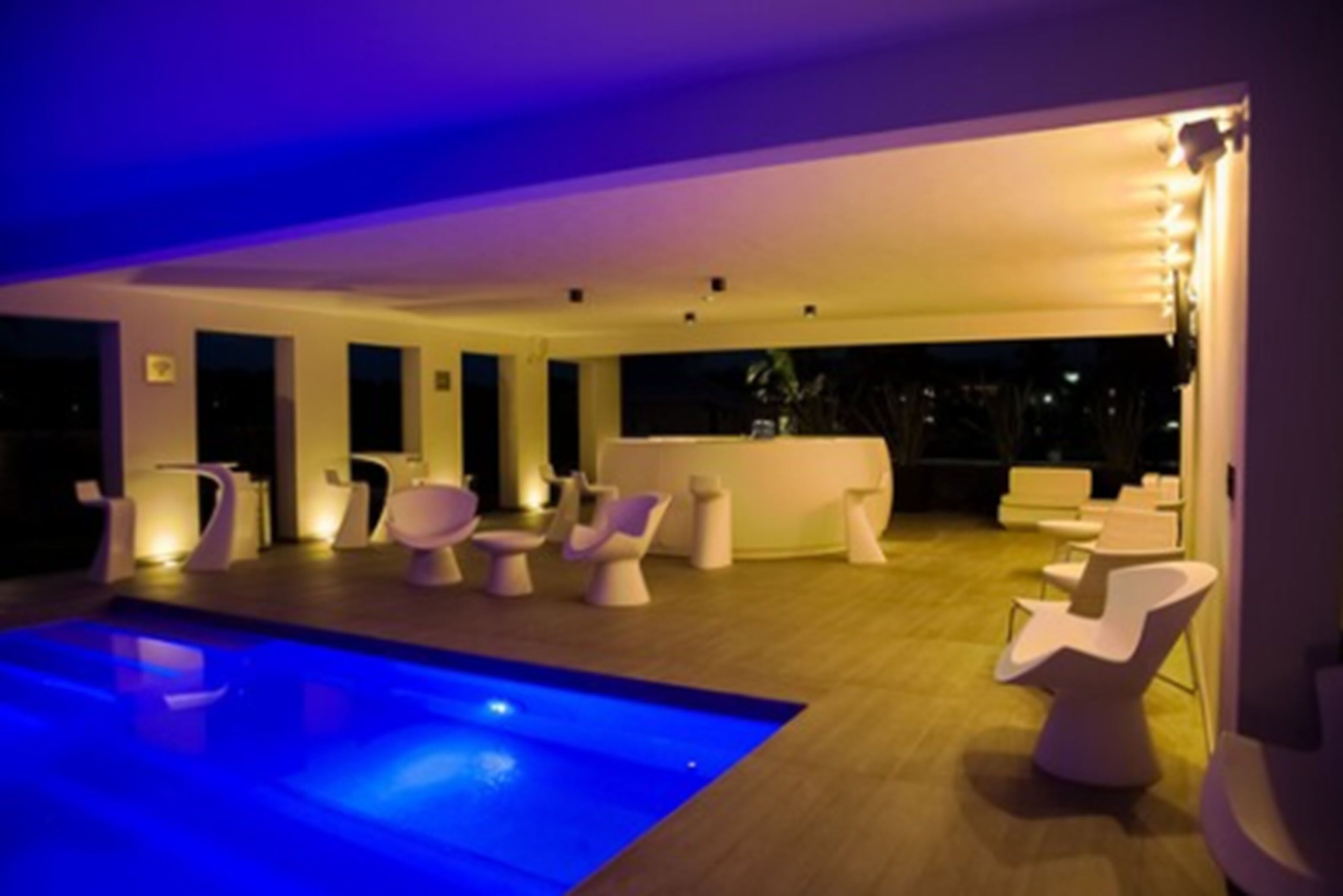 Un área interior de piscina moderna con iluminación azul, sillas blancas contemporáneas y una zona de bar con mostrador blanco.