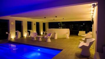 Un área interior de piscina moderna con iluminación azul, sillas blancas contemporáneas y una zona de bar con mostrador blanco.