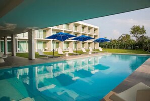 Une piscine avec des chaises longues et des parasols, un bâtiment à plusieurs étages doté de balcons, et une pelouse bien entretenue.