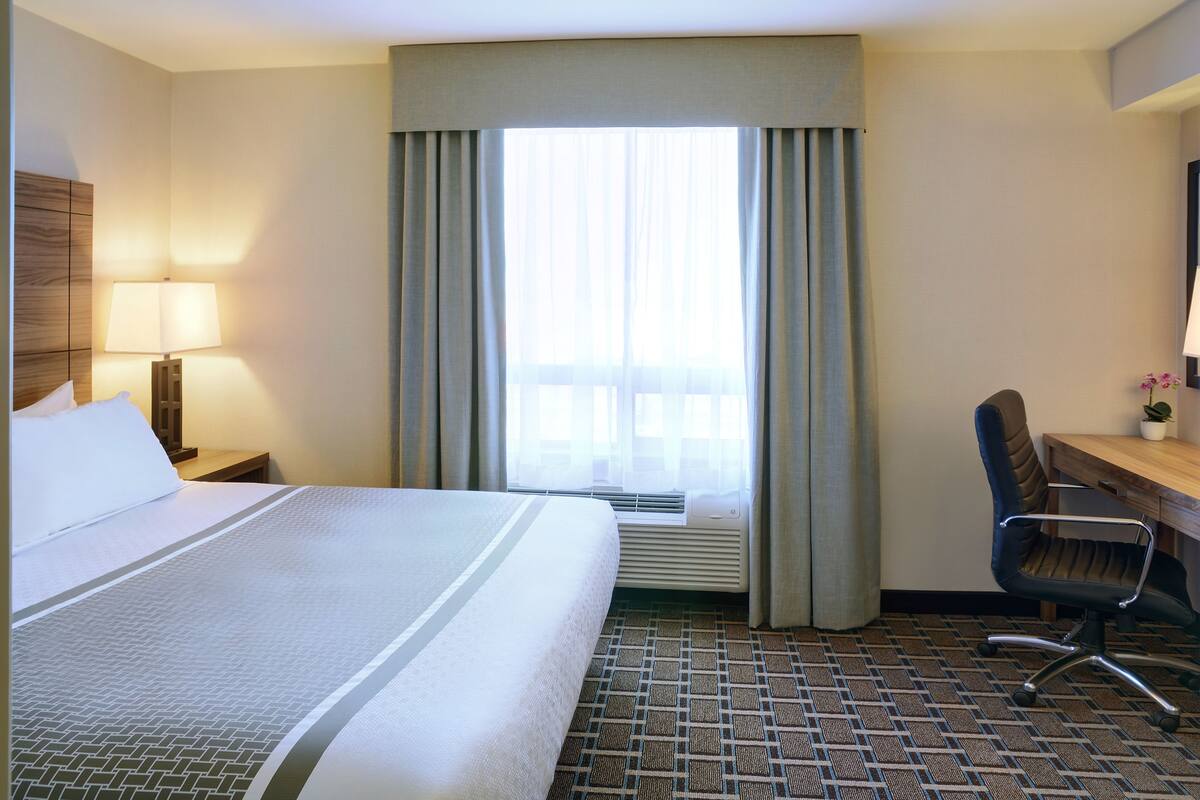 A hotel room with a large bed, a desk, a chair, a lamp, and a window with curtains.