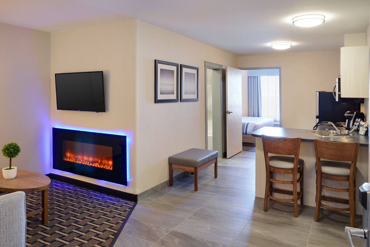 A modern hotel room with a fireplace, a flat-screen TV, a small dining area, and a kitchenette.