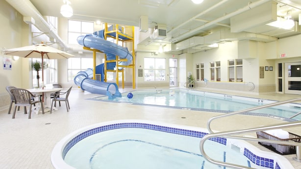 An indoor swimming pool with a blue slide, a hot tub, and a dining area with chairs and an umbrella. - Pomeroy Inn And Suites Vermilion (Vermilion)