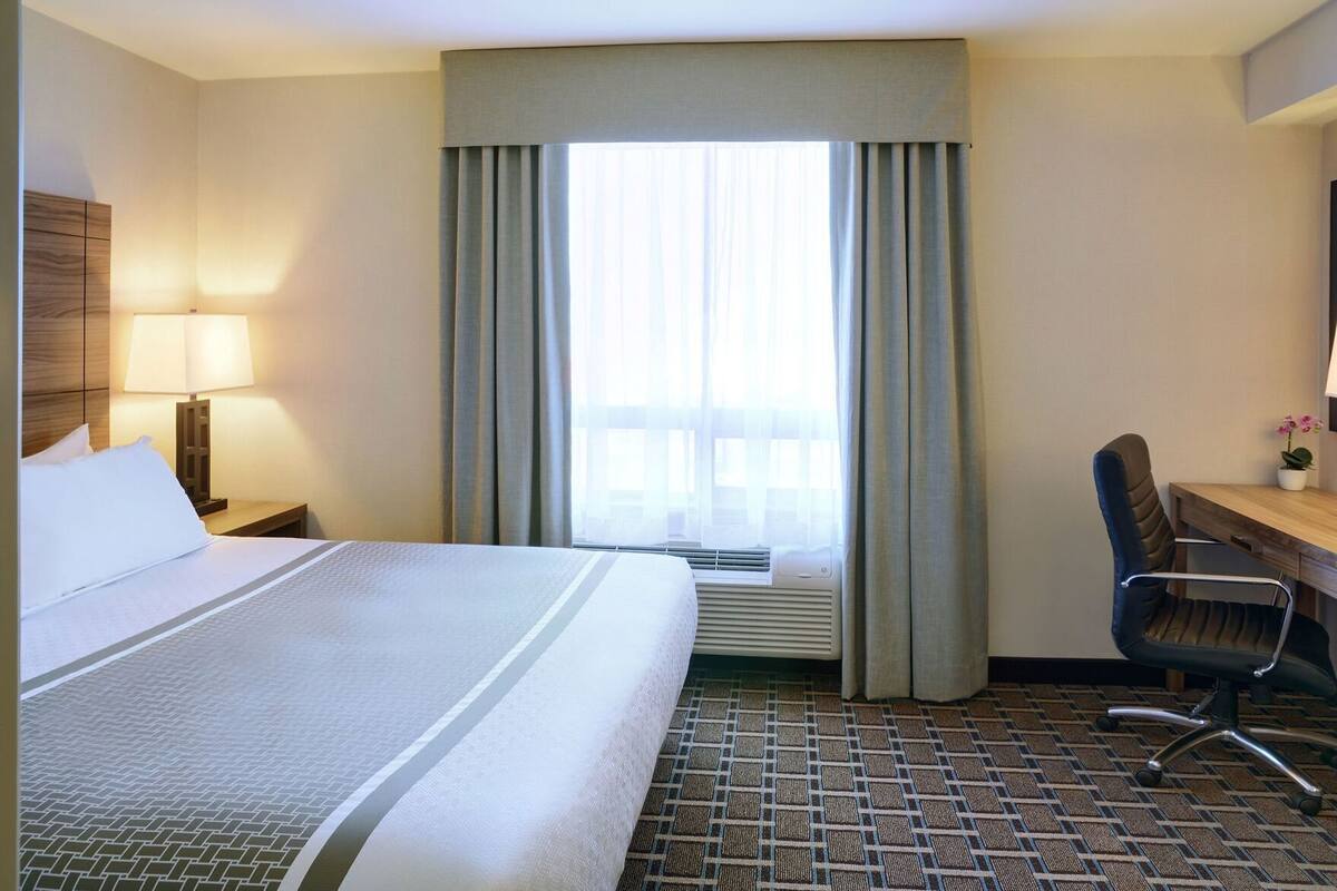 A hotel room with a bed, a desk, a chair, a lamp, and a window with curtains.