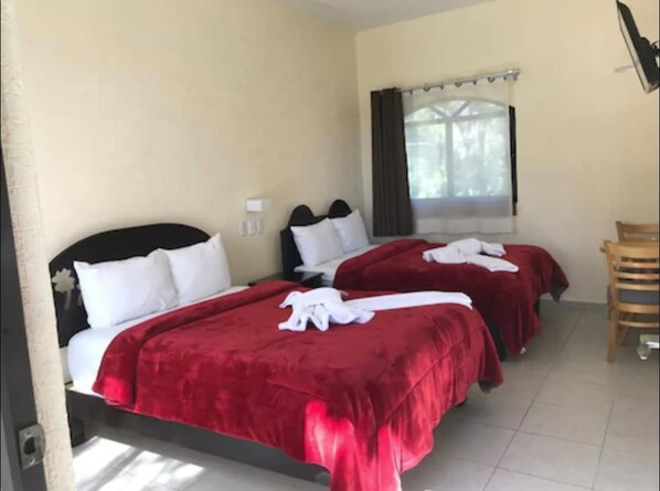 Une chambre d’hôtel avec deux lits, chacun recouvert d’une couverture rouge et doté d’oreillers blancs. On y trouve une fenêtre avec des rideaux, une chaise et un luminaire fixé au mur.