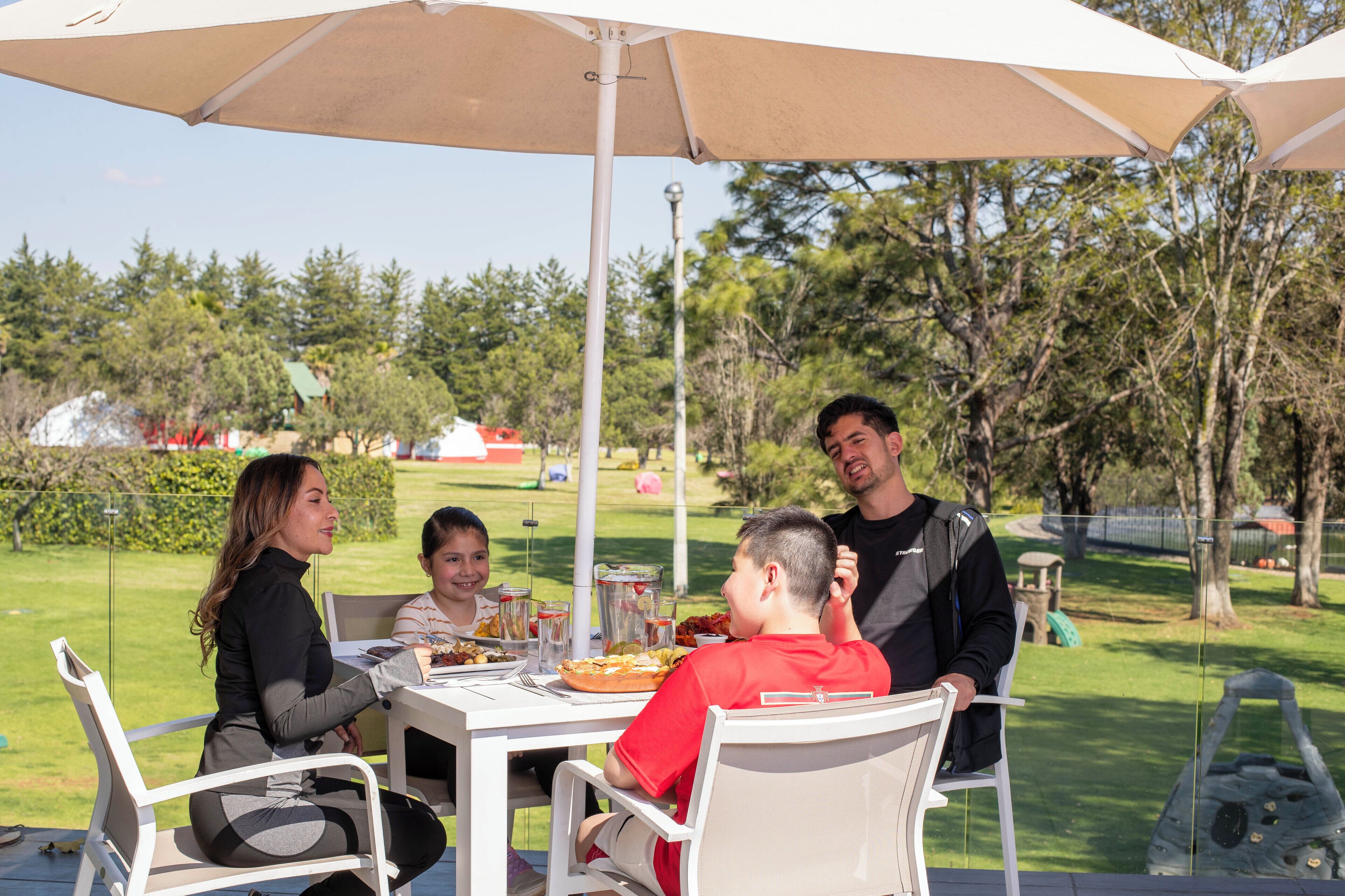 Un espace de restauration en plein air, avec une table et des chaises placées sous un grand parasol, entouré de verdure et d’arbres.