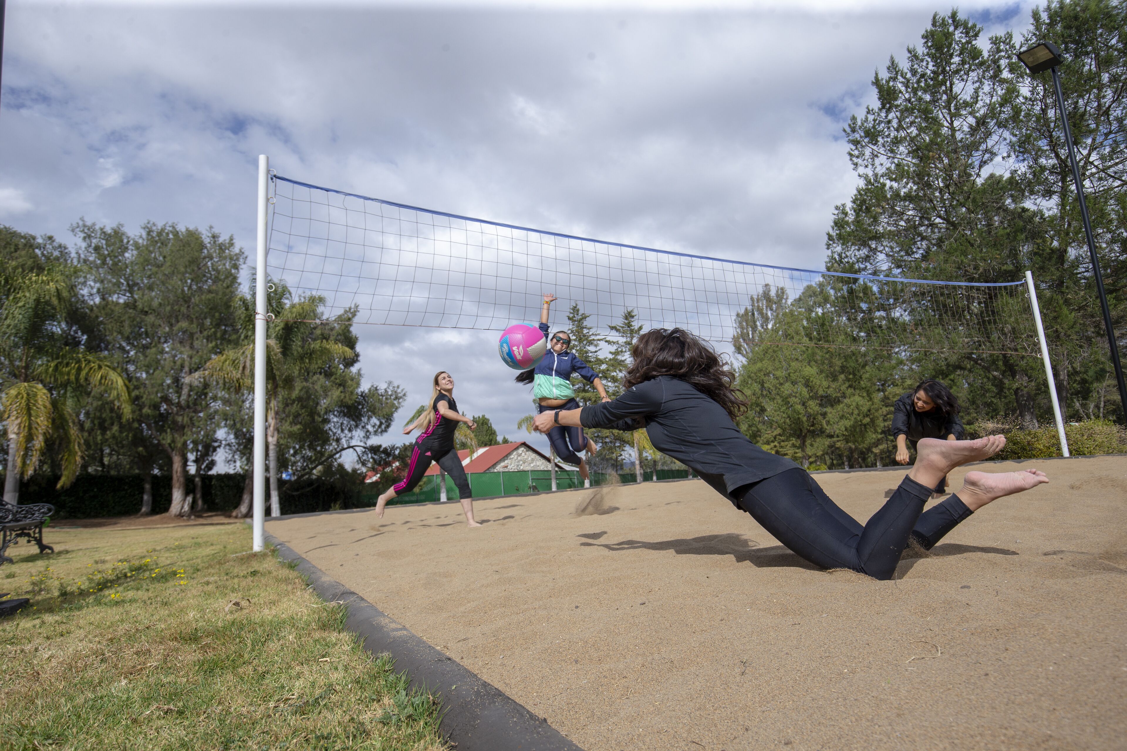 Jogo de voleibol em andamento em quadra de areia, com árvores ao fundo.