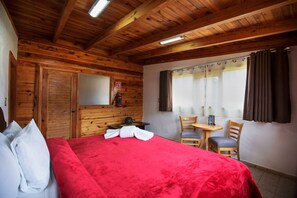 Un dormitorio de cabaña de madera con cama, una mesita, sillas y una ventana con cortinas.