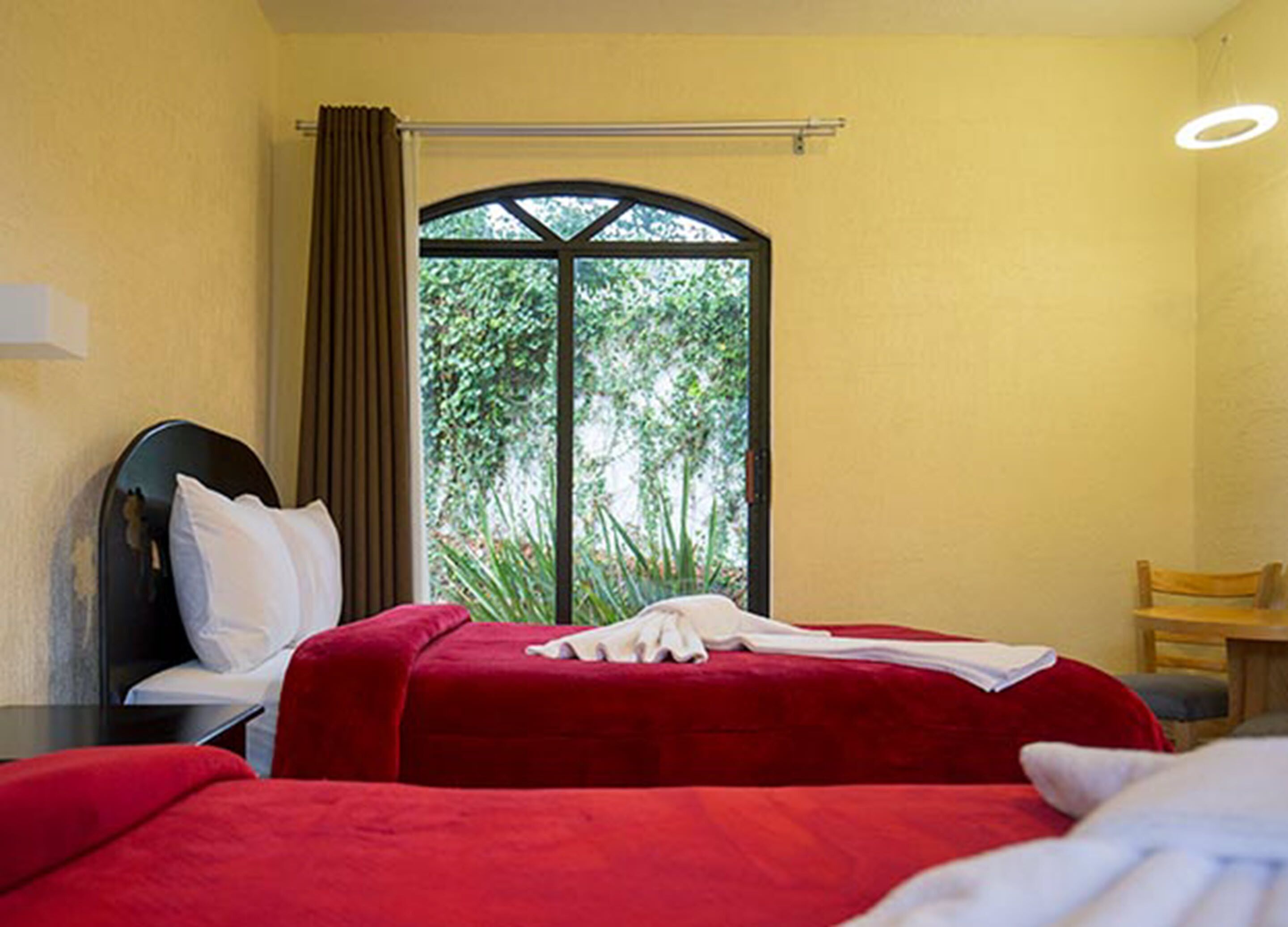 A hotel room with two beds, a window with a view of greenery, a wooden chair, and a small table.