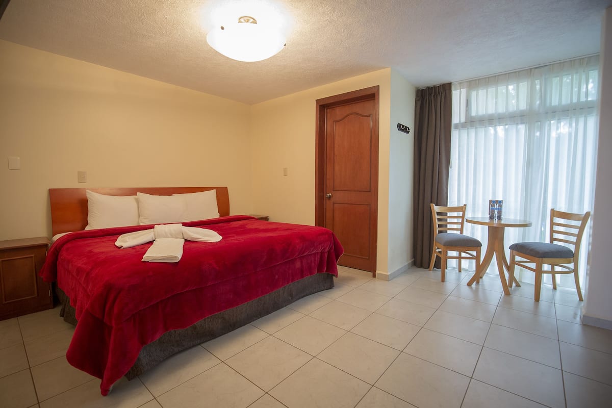 A hotel room with a bed, a wooden door, a small table with chairs, and a window with curtains.