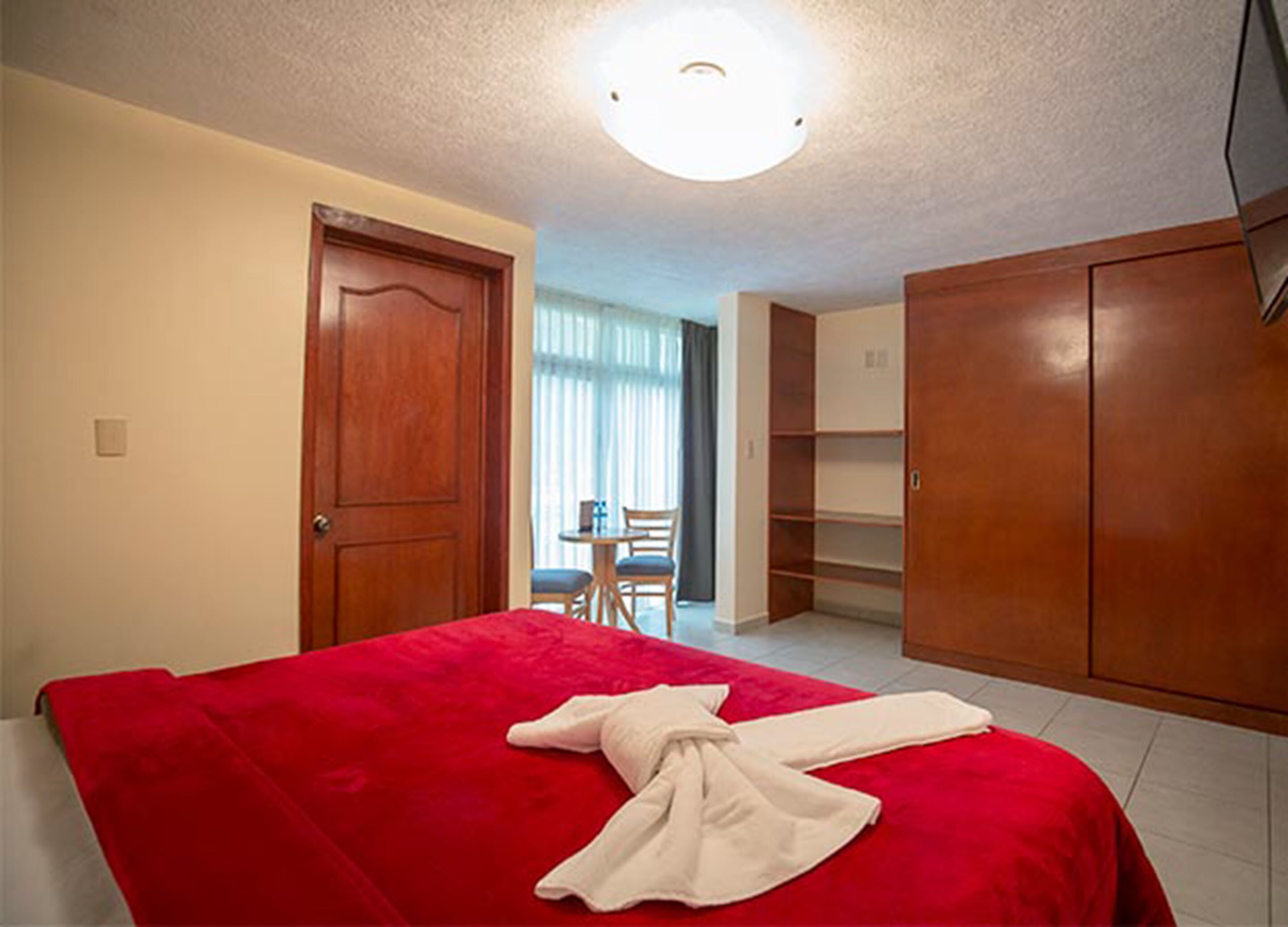 A hotel room with a red bedspread, a wooden wardrobe, and a desk with a chair.