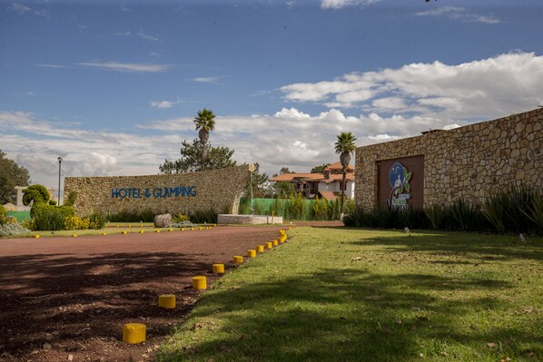 Un muro de piedra con un letrero que dice "HOTEL & GLAMPING" y un sendero que conduce a un edificio con techo rojo.