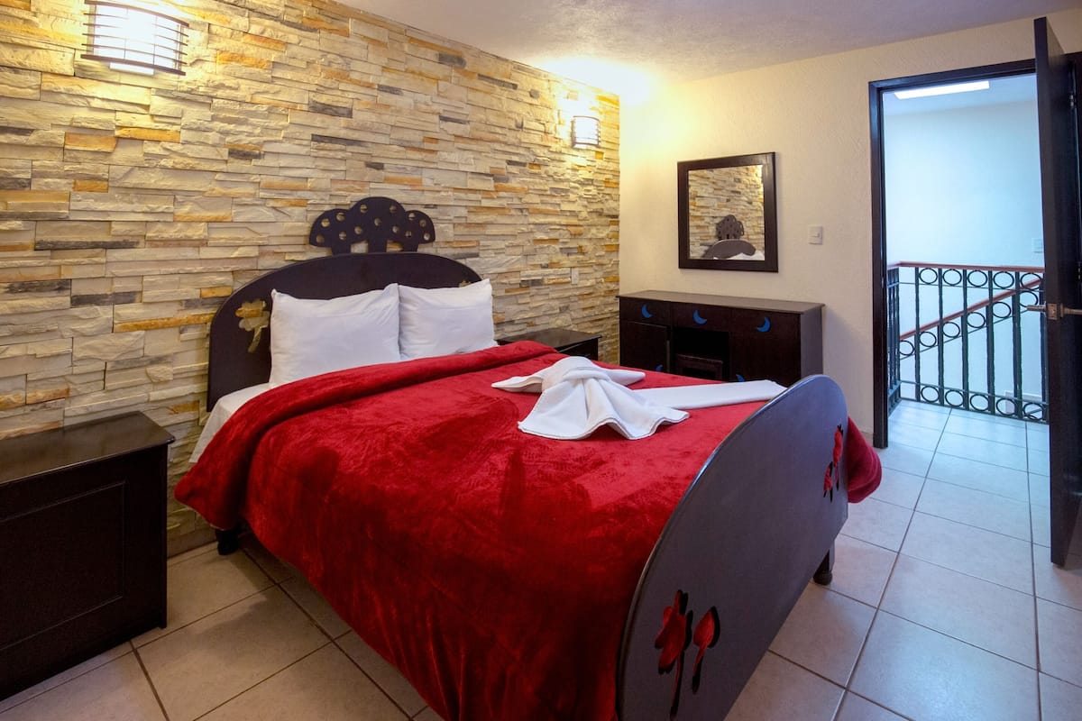 A bedroom with a stone wall, a bed with a red blanket, a nightstand, a dresser, and a door leading to a balcony.