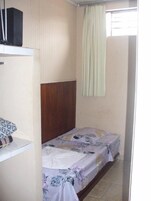 Una habitación pequeña con una cama individual, cabecera de madera, ropa de cama estampada con motivos florales, un armario blanco y una ventana con cortina.