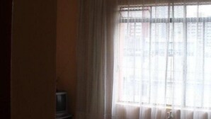 Una habitación con cama, ventilador de techo, ventana con cortinas traslúcidas y una pequeña mesa con televisor.