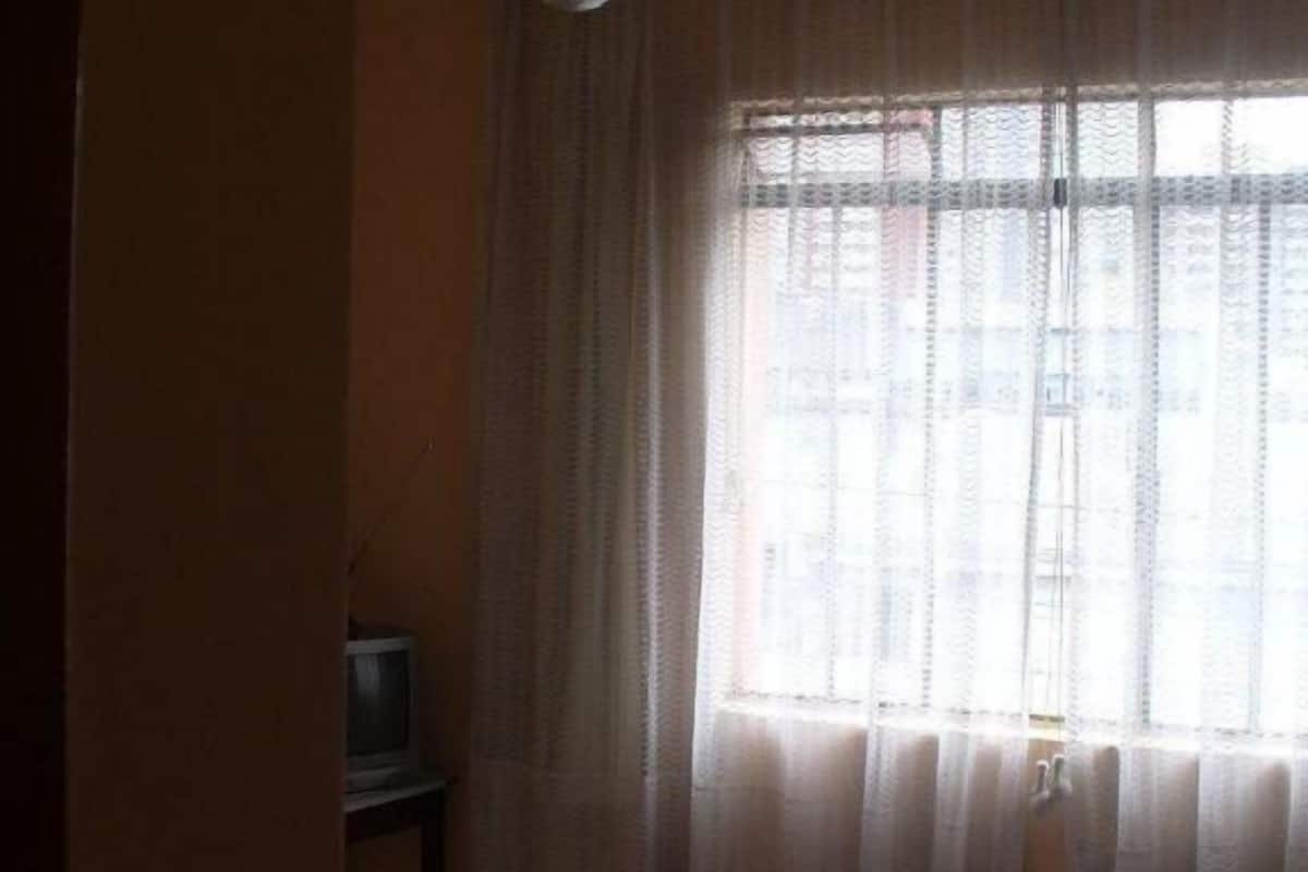 Una habitación con cama, ventilador de techo, ventana con cortinas traslúcidas y una pequeña mesa con televisor.