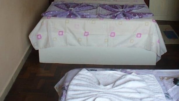Una cama individual con cabecera de madera, una colcha blanca con detalles morados y un cojín a juego.