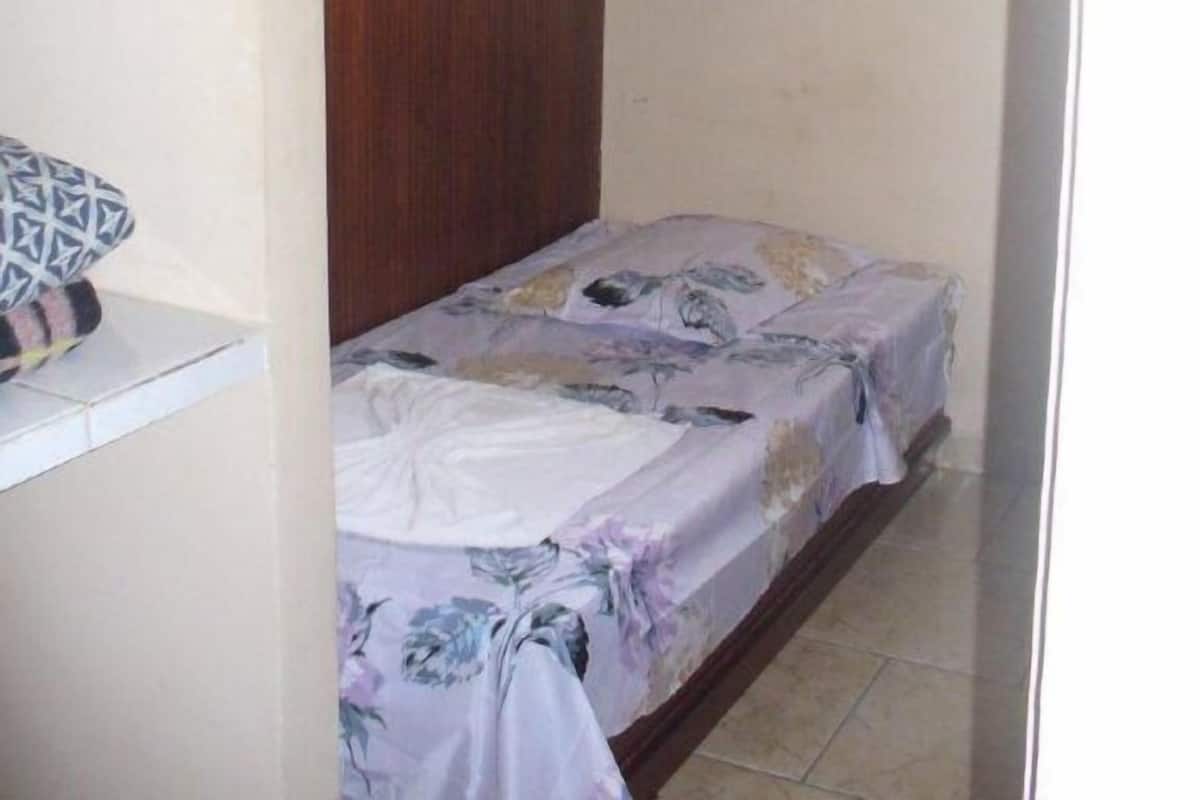 Una habitación pequeña con una cama individual, cabecera de madera, armario blanco y ventana con cortina.