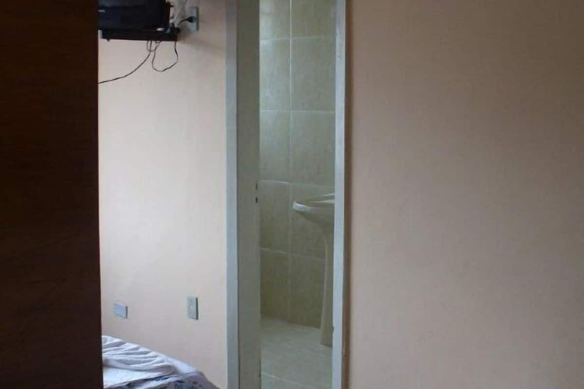 Una habitación pequeña y escasamente amoblada, con dos camas, un televisor en la pared y una zona de ducha visible a través de una puerta de vidrio.