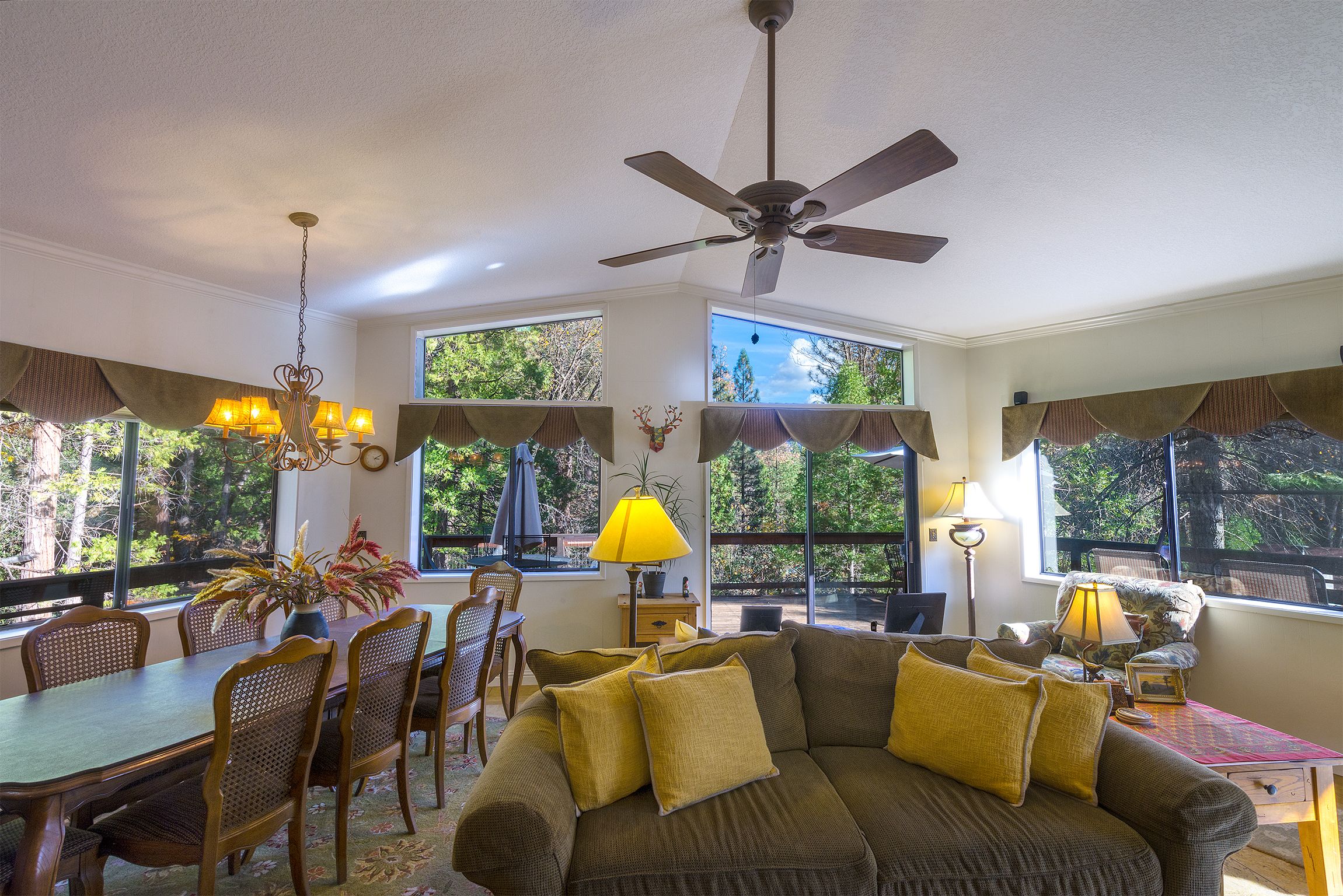 A dining area with a table and chairs, a sofa with cushions, a ceiling fan, and large windows with curtains.