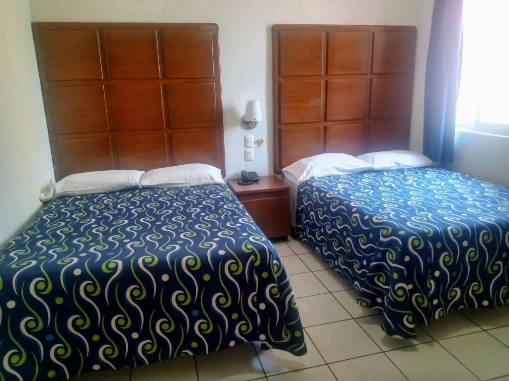 A hotel room with two beds, each with a patterned bedspread, a wooden headboard, a bedside table with a lamp, and a window with curtains.