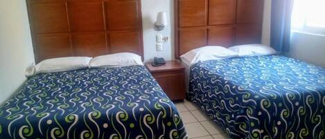 A hotel room with two beds, each with a patterned bedspread, a wooden headboard, a bedside table with a lamp, and a window with curtains.