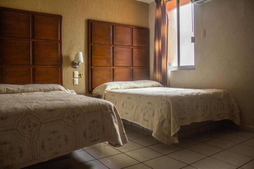 A hotel room with two beds, a wooden headboard, a wall-mounted lamp, and a window with curtains.