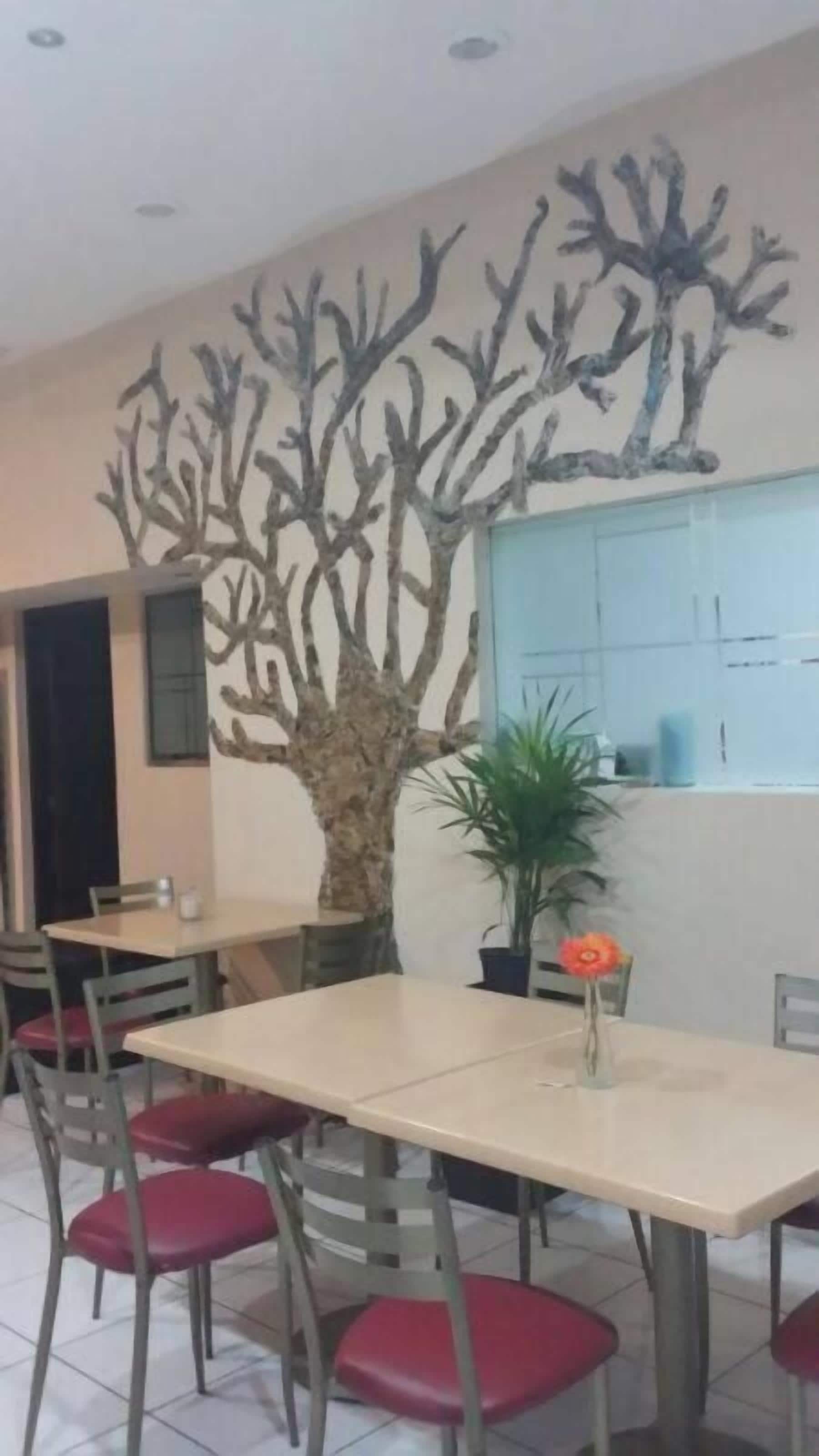 Un área de comedor con mesas y sillas, una pared decorativa con estructuras que recuerdan a árboles, y una maceta con una flor roja.