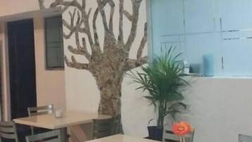 Un área de comedor con mesas y sillas, una pared decorativa con estructuras que recuerdan a árboles, y una maceta con una flor roja.
