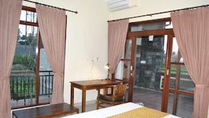A bedroom with a bed, a desk, and a chair. There is a window with curtains, a lamp, and a view of the outside. - Ganesha Home Stay (Ubud)