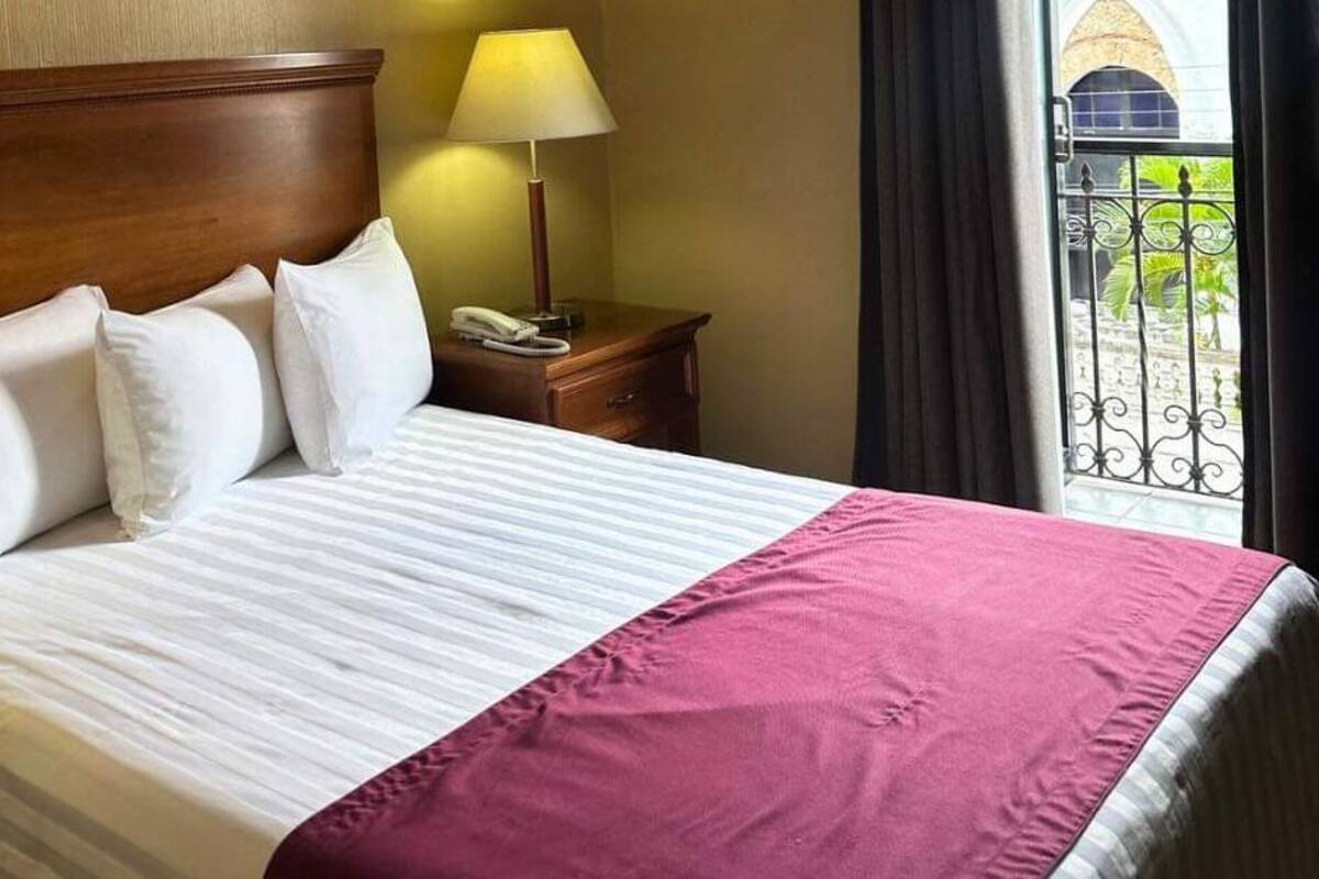 A neatly made bed with white and pink linens, a wooden headboard, a bedside lamp, and a view of a balcony with a wrought iron railing.