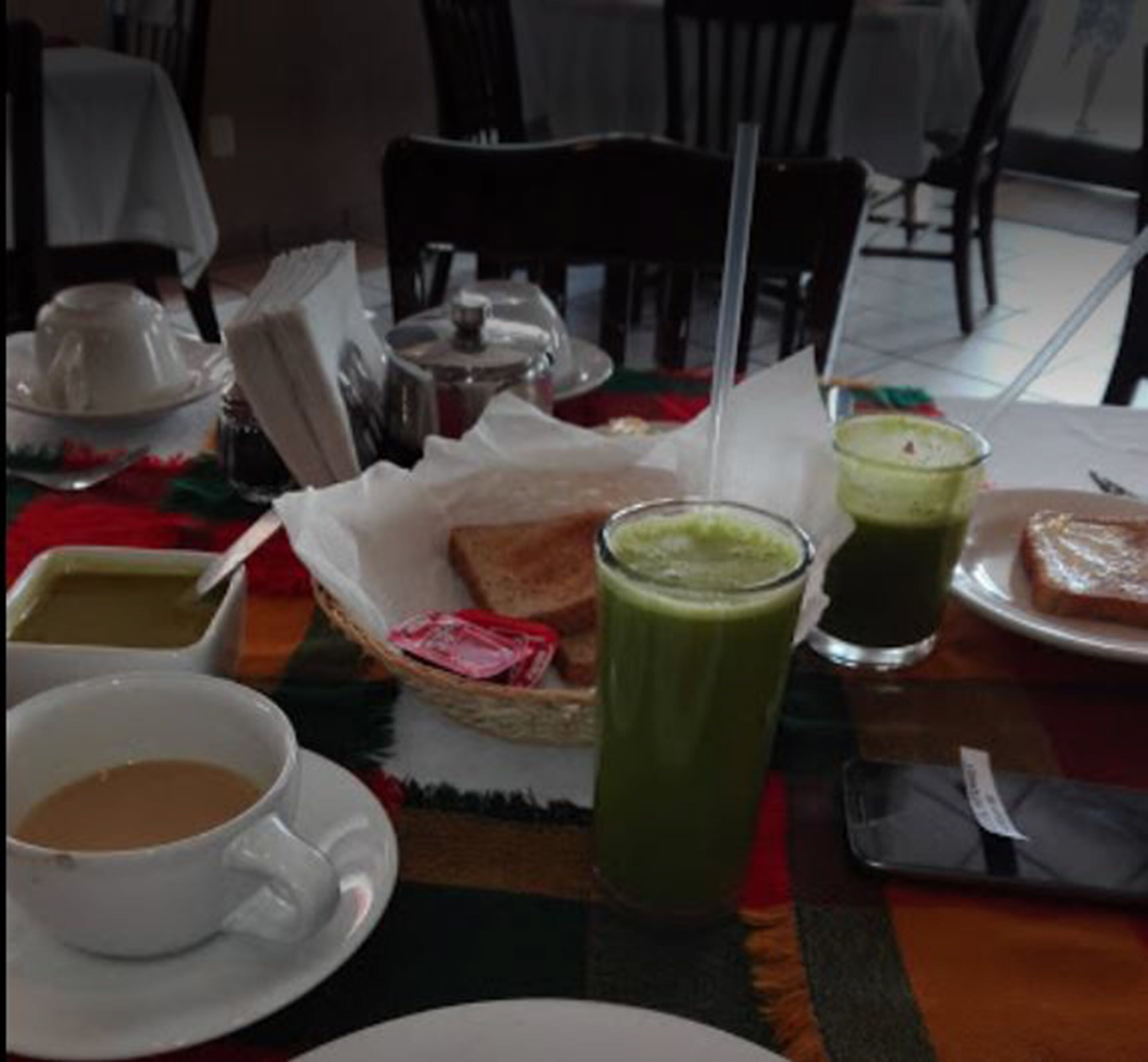 A table set with a teapot, cups, a basket of bread, a green smoothie, and a plate of toast.