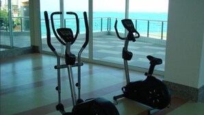 Een overdekte fitnessruimte met twee hometrainers en een raam met uitzicht op een balkon en de zee.