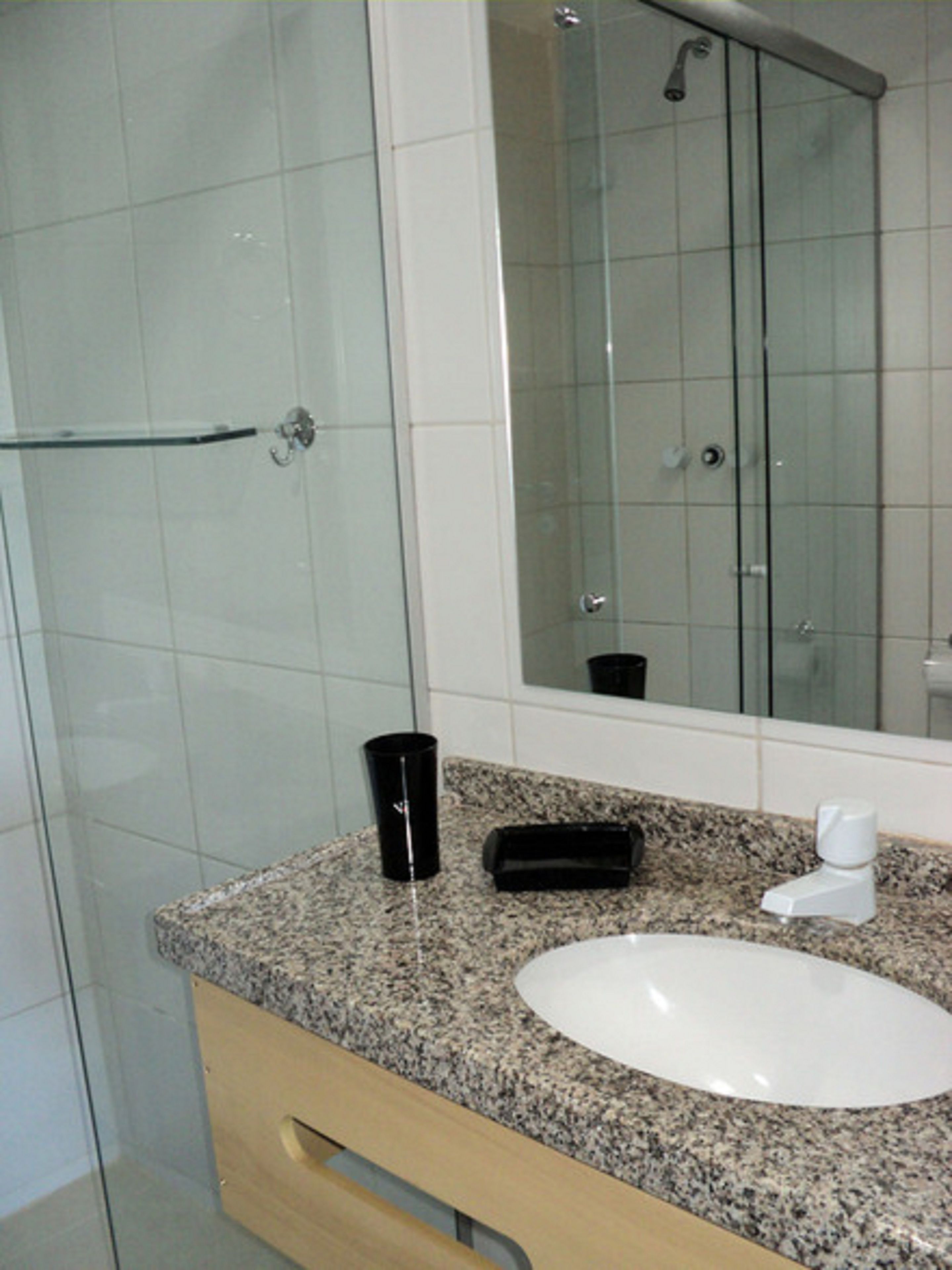Un baño con cabina de ducha de vidrio, un lavabo blanco, una taza negra y un espejo.