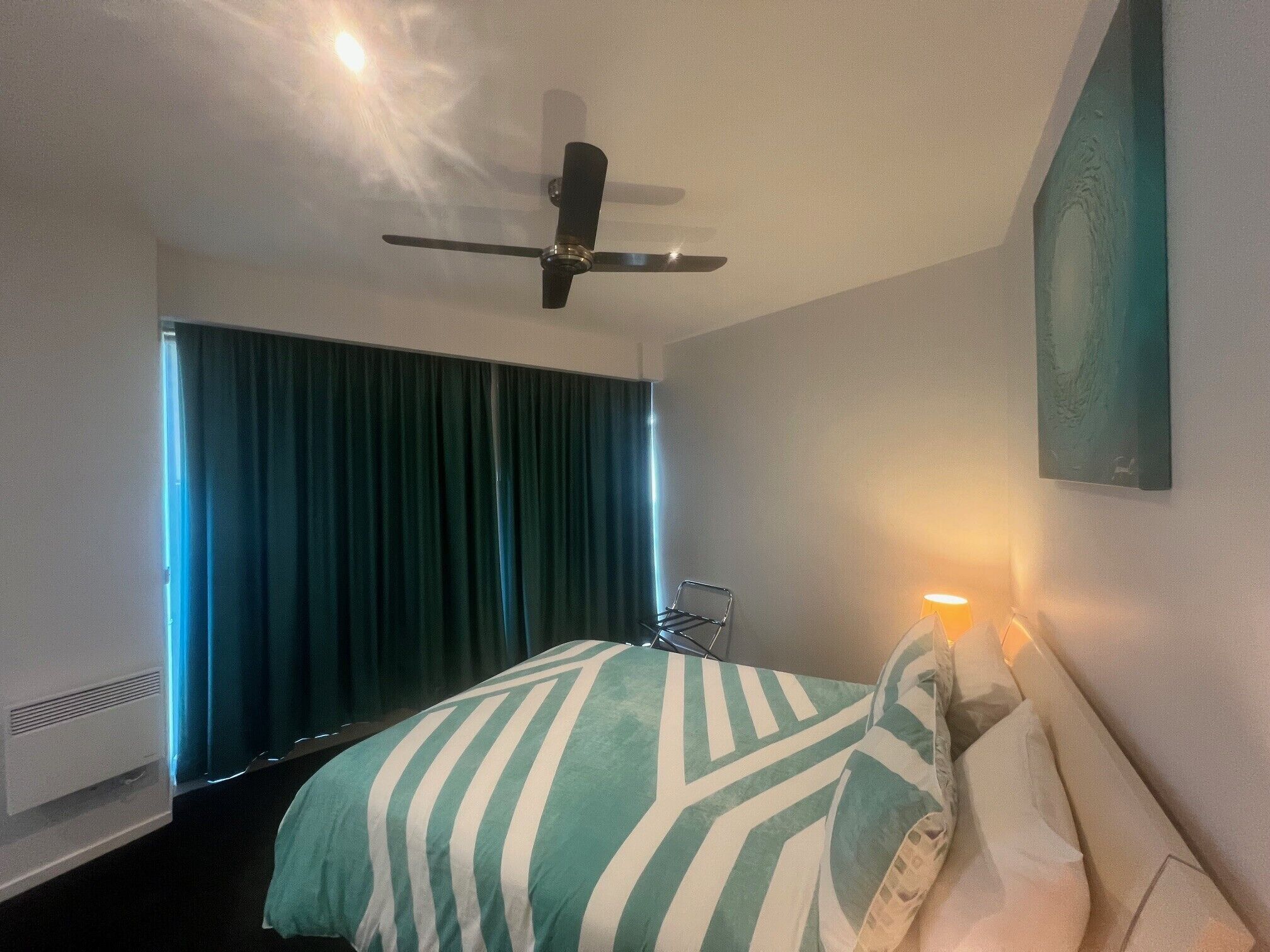 A hotel room with a bed, a ceiling fan, a window with curtains, and a wall-mounted artwork.