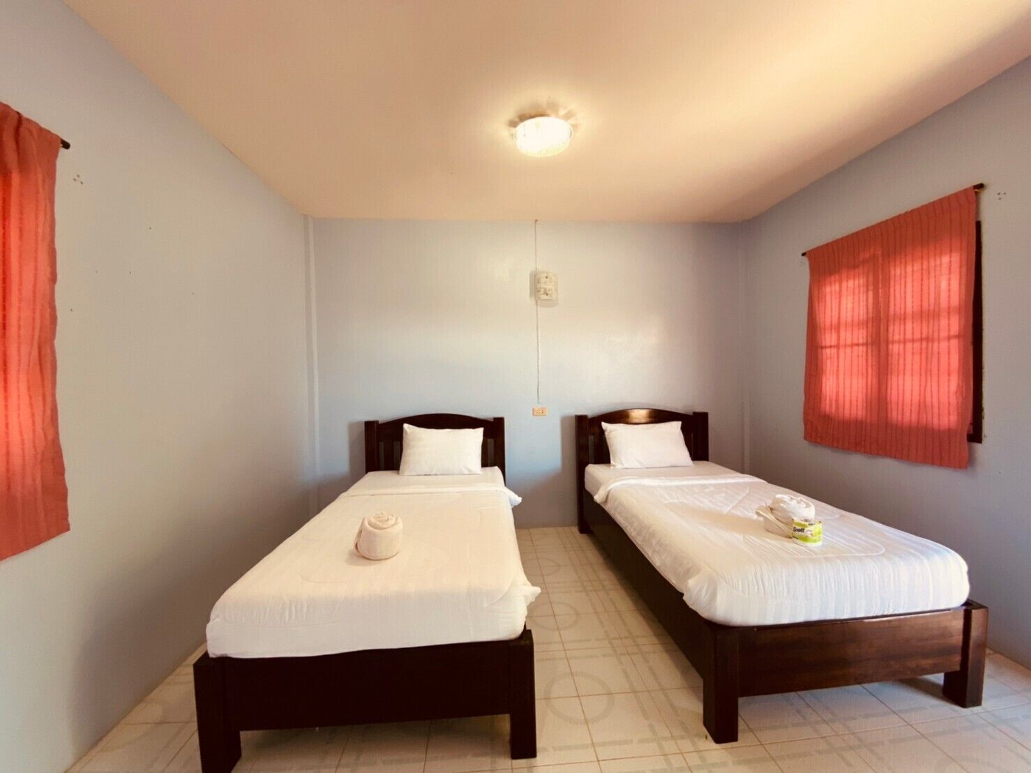 A room with two single beds, each with a wooden headboard and white bedding. There is a small table between the beds, and the room has a tiled floor.