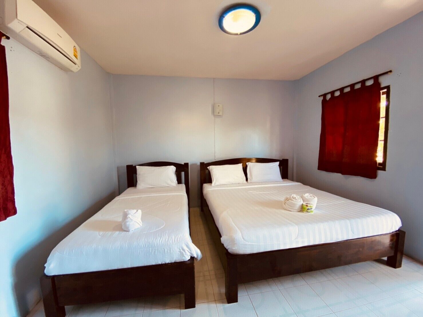 A hotel room with two single beds, a ceiling light, an air conditioning unit, and a window with red curtains.