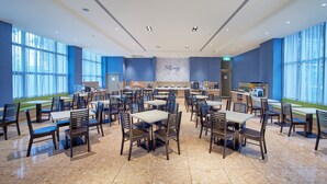 Uma sala de jantar ampla com várias mesas e cadeiras, área de cozinha e grandes janelas que permitem entrada de luz natural.