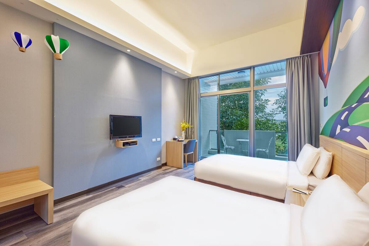 Ein Hotelzimmer mit zwei Betten, einem Schreibtisch, einem Fernseher und einem Balkon mit Blick auf Grünflächen.