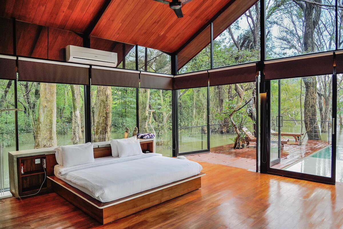 A spacious bedroom with a large bed, wooden flooring, and a view of a forested area through large windows.
