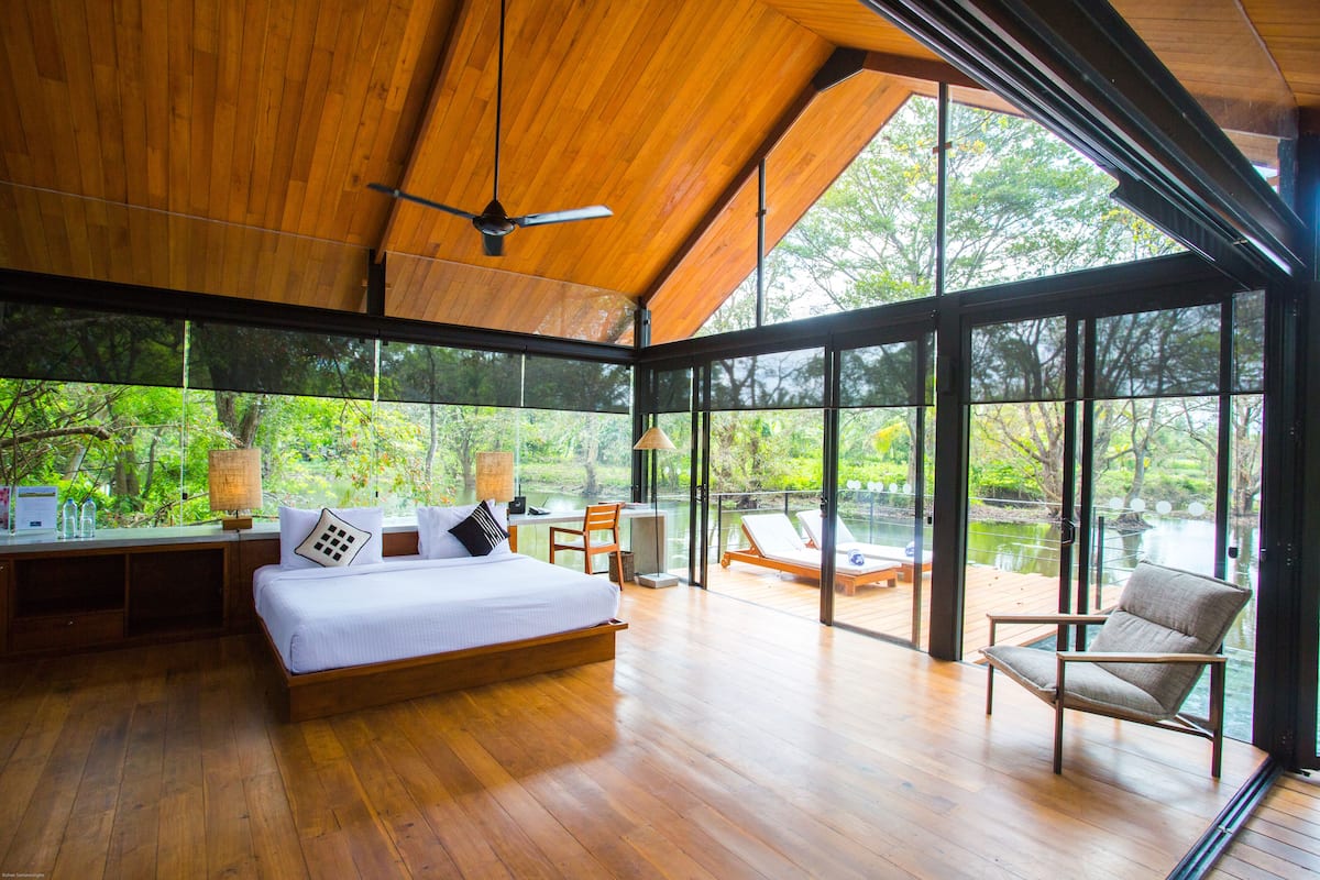 A spacious bedroom with a large bed, a desk, and a chair, overlooking a lake through large windows.